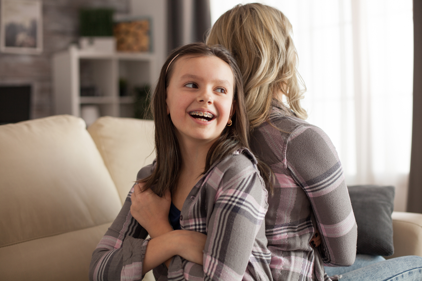 Smiling girl with braces sitting next to a woman, both in plaid shirts, in a cozy living room setting, emphasizing comfort and connection before a dental cleaning appointment.